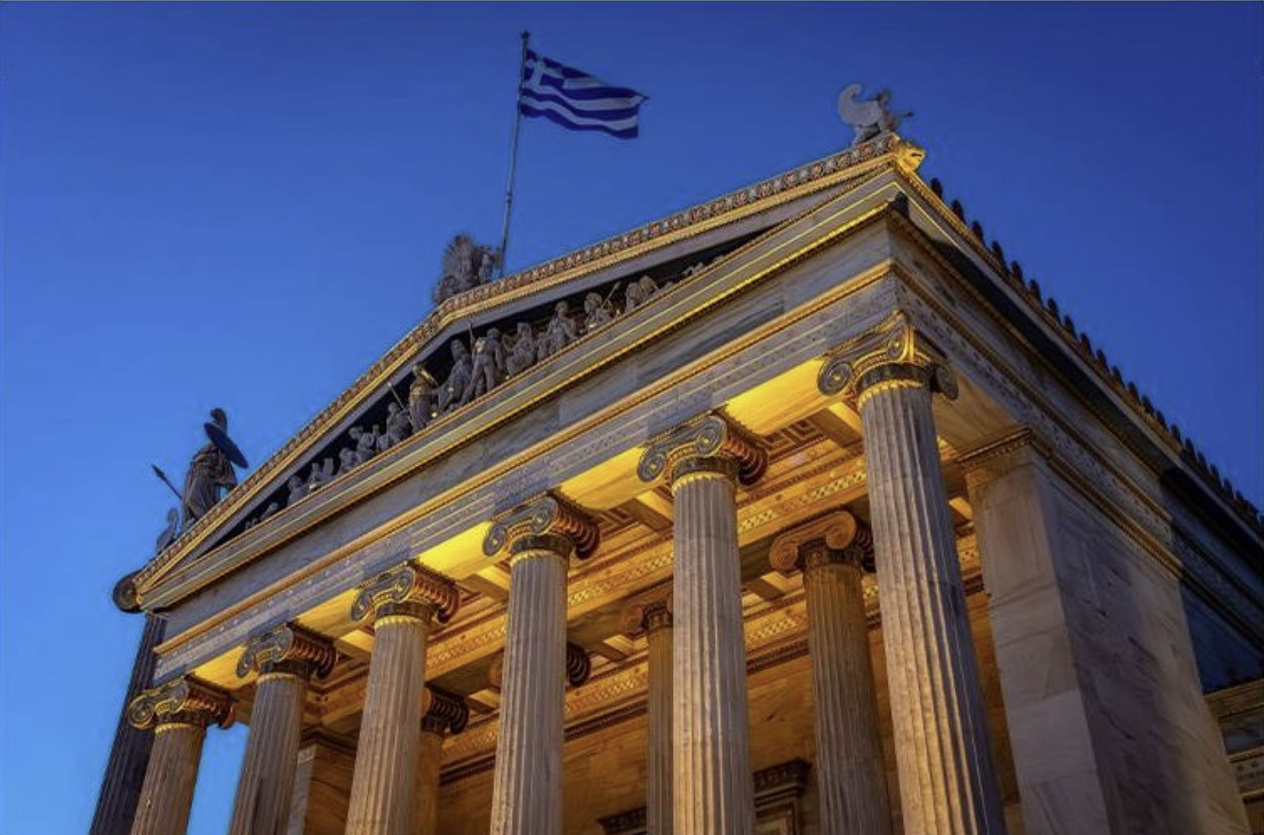 Η πρόσοψη του Μεγάρου της Ακαδημίας Αθηνών φωτισμένη το σούρουπο, με την ελληνική σημαία